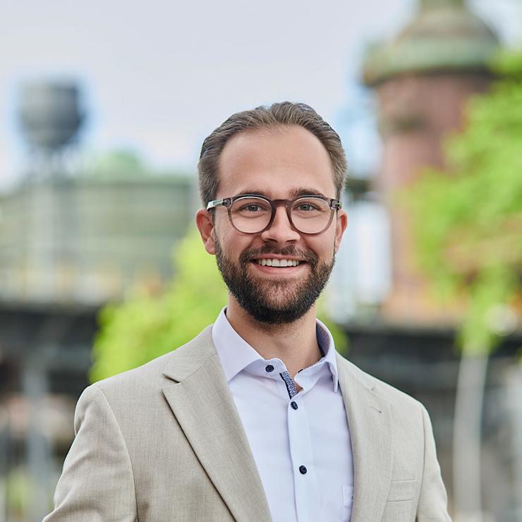 Mann mit Brille und weißem Hemd steht in einem hellen Flur mit Pflanzen im Hintergrund.