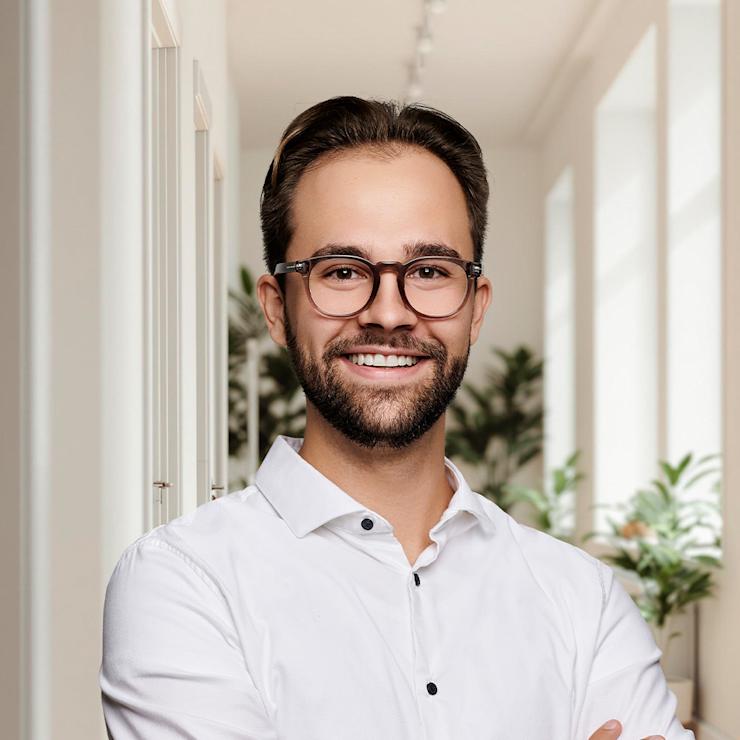 Mann mit Brille und weißem Hemd steht in einem hellen Flur mit Pflanzen im Hintergrund.