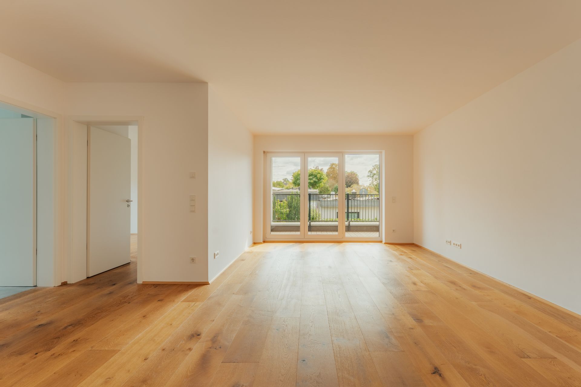 Helles, leeres Wohnzimmer mit Holzboden und großen Fenstern zum Balkon.