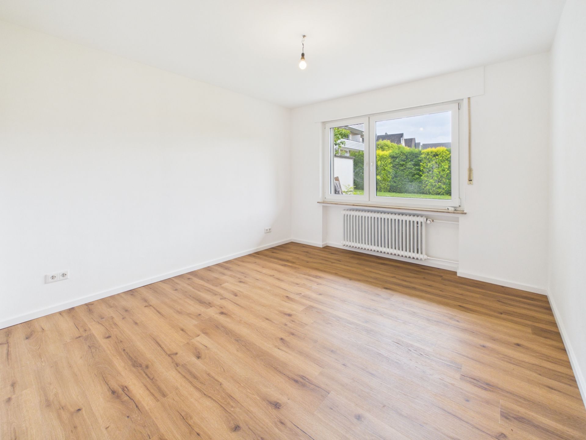 Leerer Raum mit Holzfußboden, weiß gestrichenen Wänden und großem Fenster mit Blick ins Grüne.