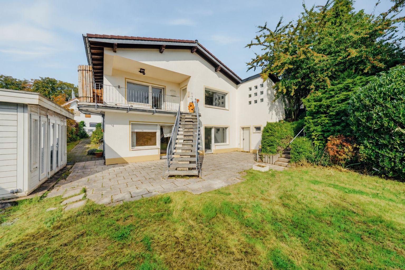 Zweistöckiges Wohnhaus mit Außentreppe, Garten und Terrasse, umgeben von Büschen und Bäumen.