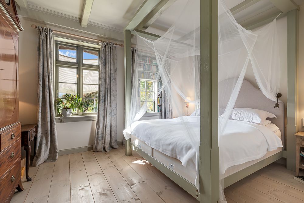 Helles Schlafzimmer mit Himmelbett, Moskitonetz und zwei Fenstern mit Vorhängen.