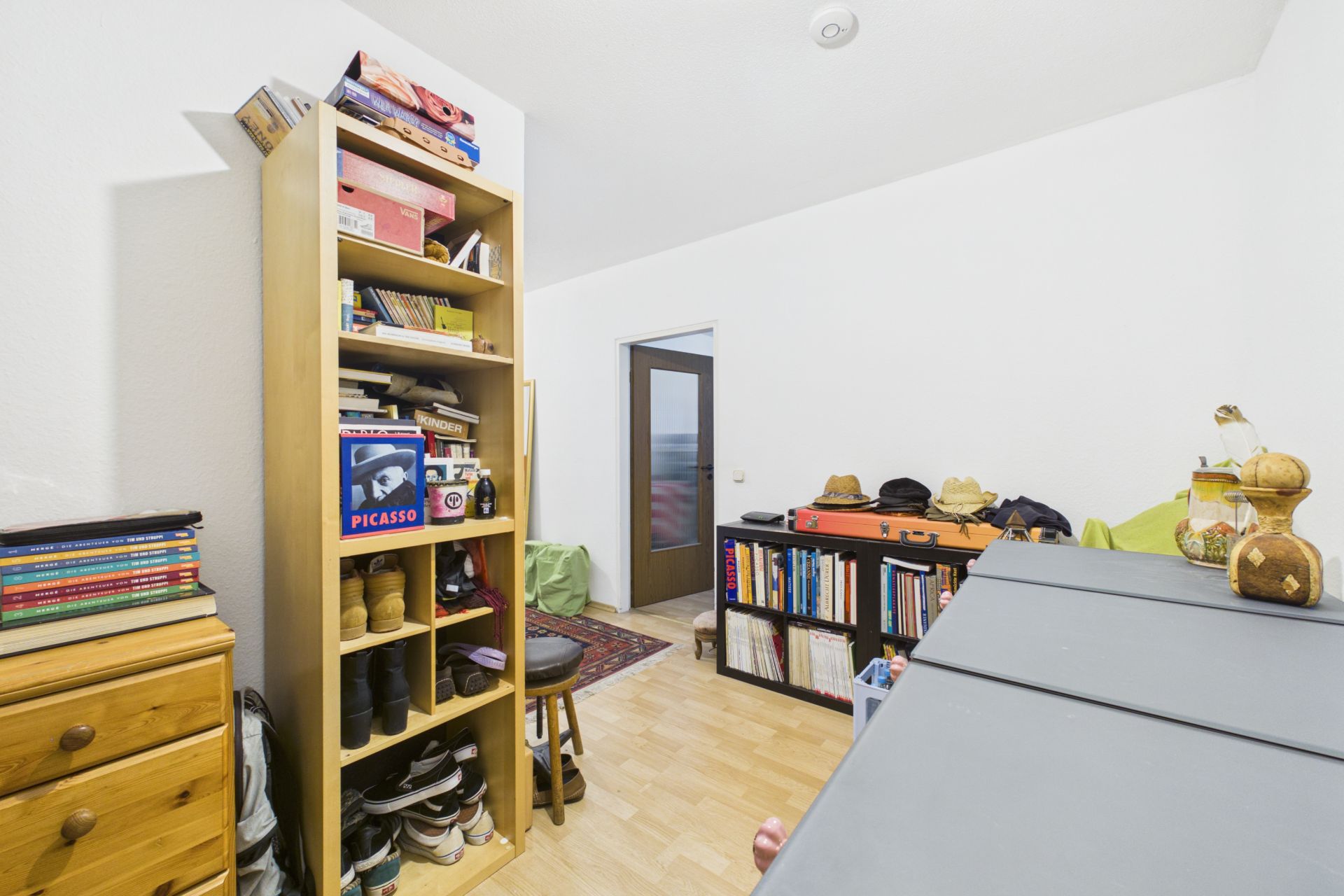 Kleines Zimmer mit Bücherregalen, Schuhregal, Kommode und dekorativen Gegenständen.