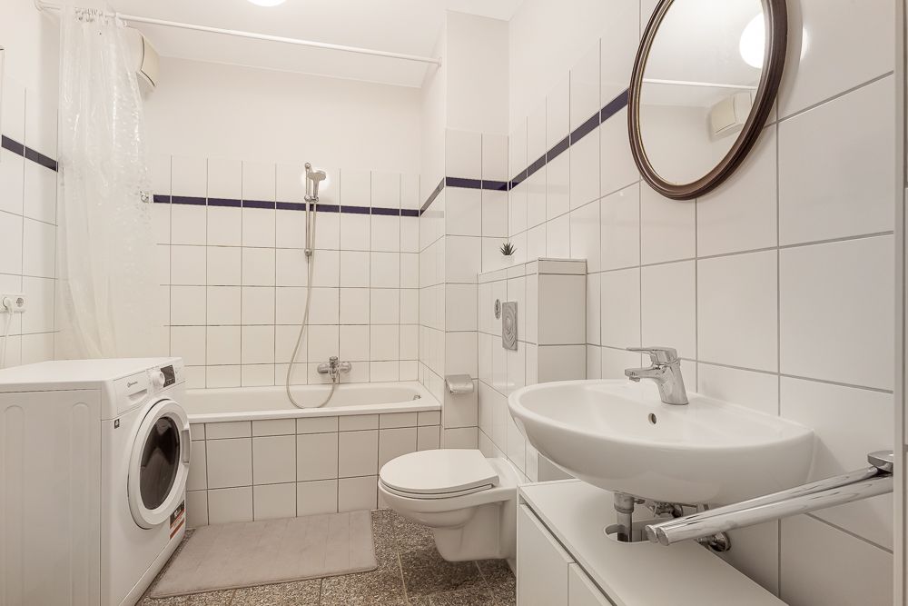 Helles Badezimmer mit Badewanne, Waschmaschine, WC, Waschbecken und rundem Spiegel.