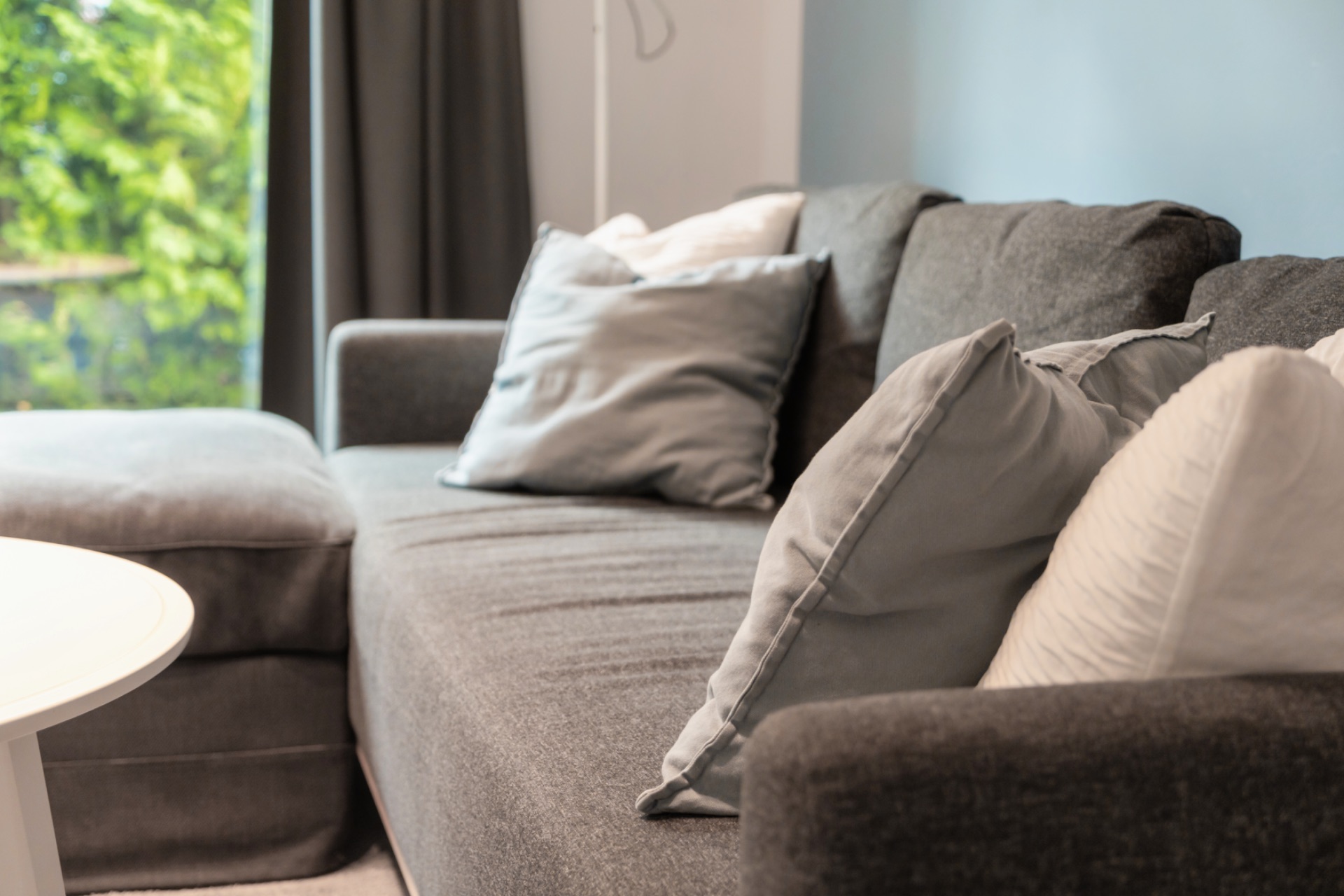 Graues Sofa mit mehreren Kissen vor einem Fenster mit Vorhang, daneben ein runder Tisch.