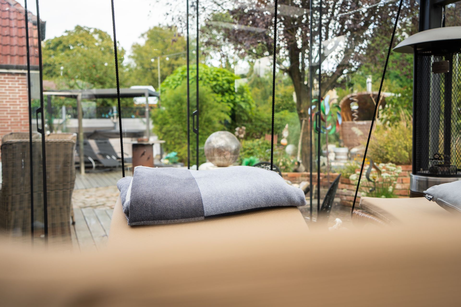 Blick durch Glas auf Garten mit Bäumen, Terrasse und einer grauen Decke im Vordergrund.