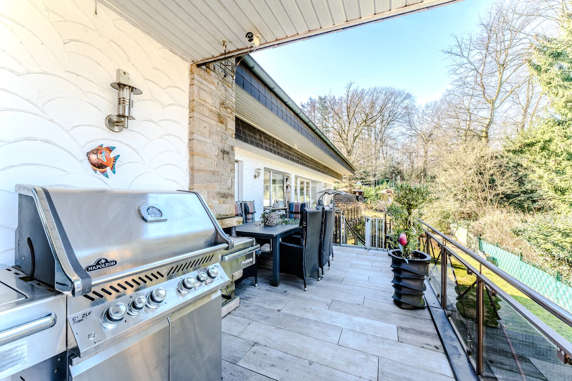 Große Terrasse mit Grill, Esstisch und Stühlen, umgeben von Bäumen und Garten.
