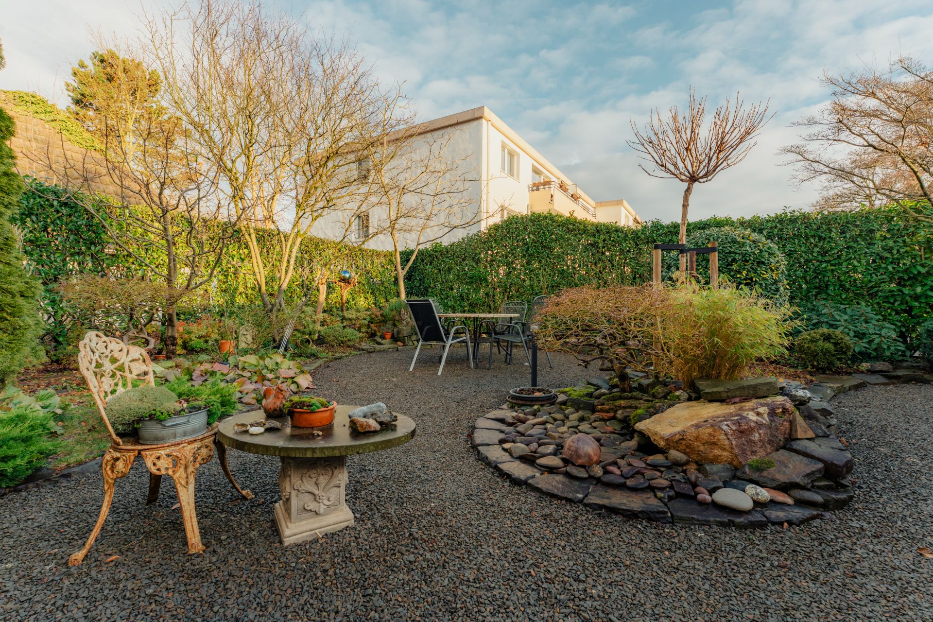 Garten mit Kiesweg, runder Steintisch, Stühlen und kleinem Teich, umgeben von Büschen und Bäumen.