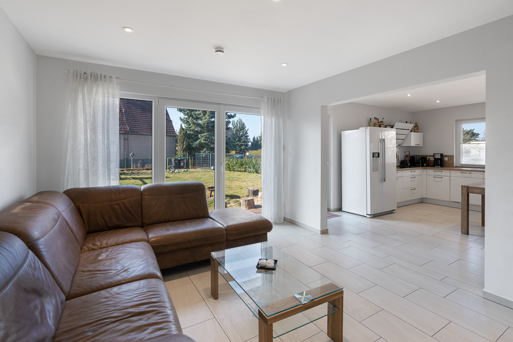 Wohnzimmer mit braunem Ecksofa, Glastisch und Blick in offene Küche mit großem Fenster.