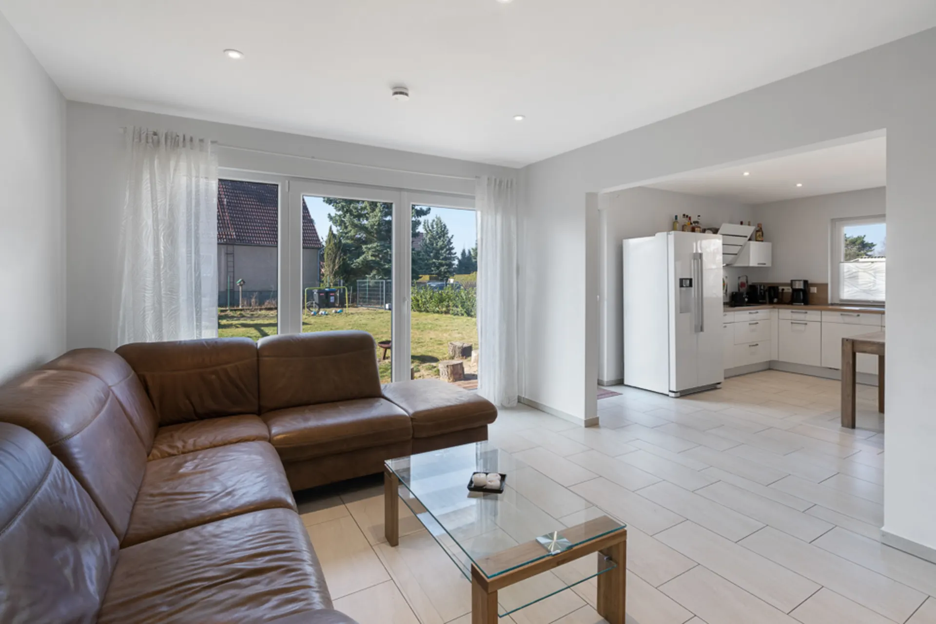 Wohnzimmer mit braunem Ecksofa, Glastisch und Blick in offene Küche mit großem Fenster.