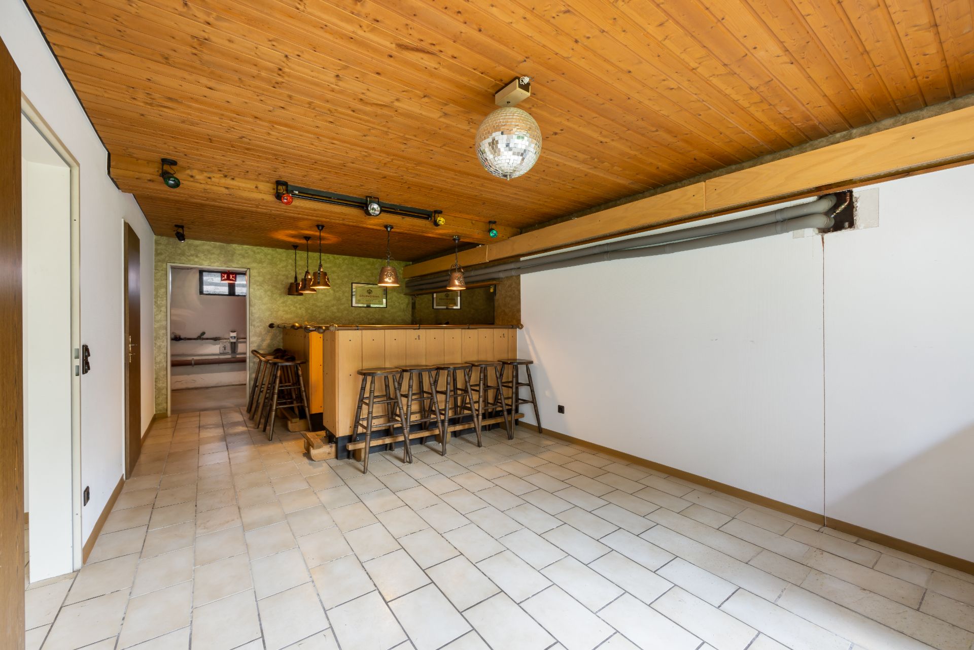 Raum mit Holzdecke, Bartresen und mehreren Barhockern auf gefliestem Boden.
