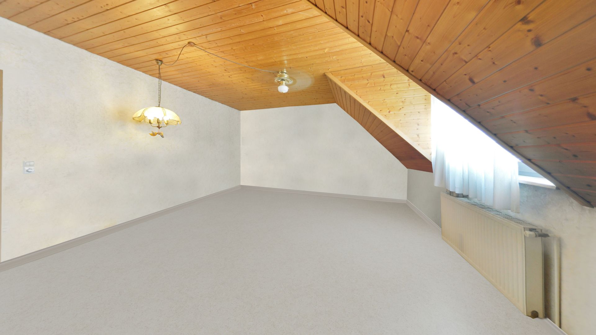 Leeres Dachzimmer mit schräger Holzdecke, Teppichboden, Fenster und Heizkörper.
