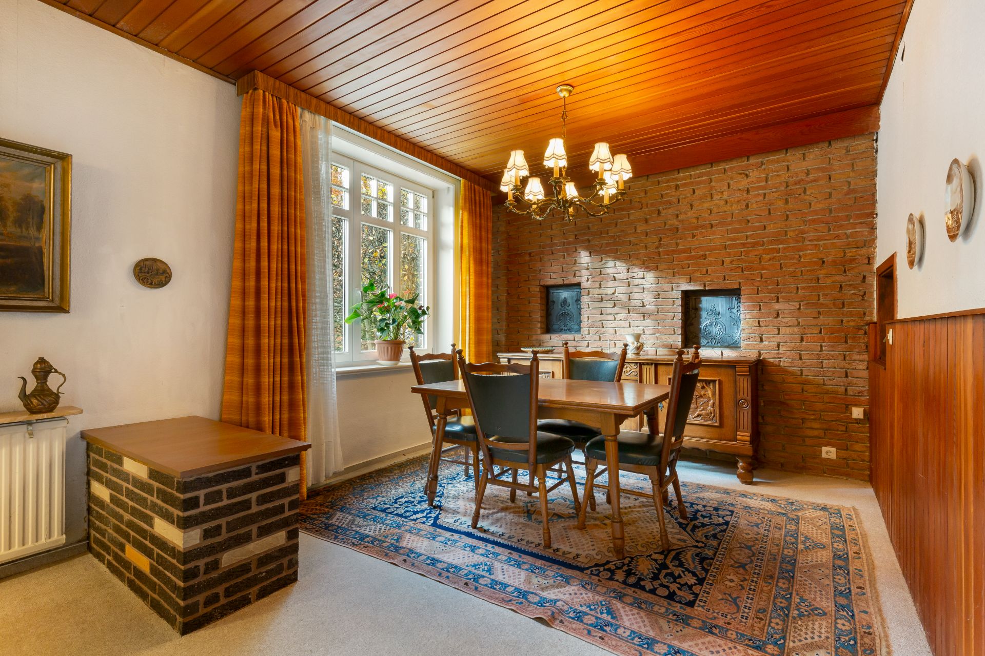 Esszimmer mit Holztisch, sechs Stühlen, Teppich, Kronleuchter und Backsteinwand.