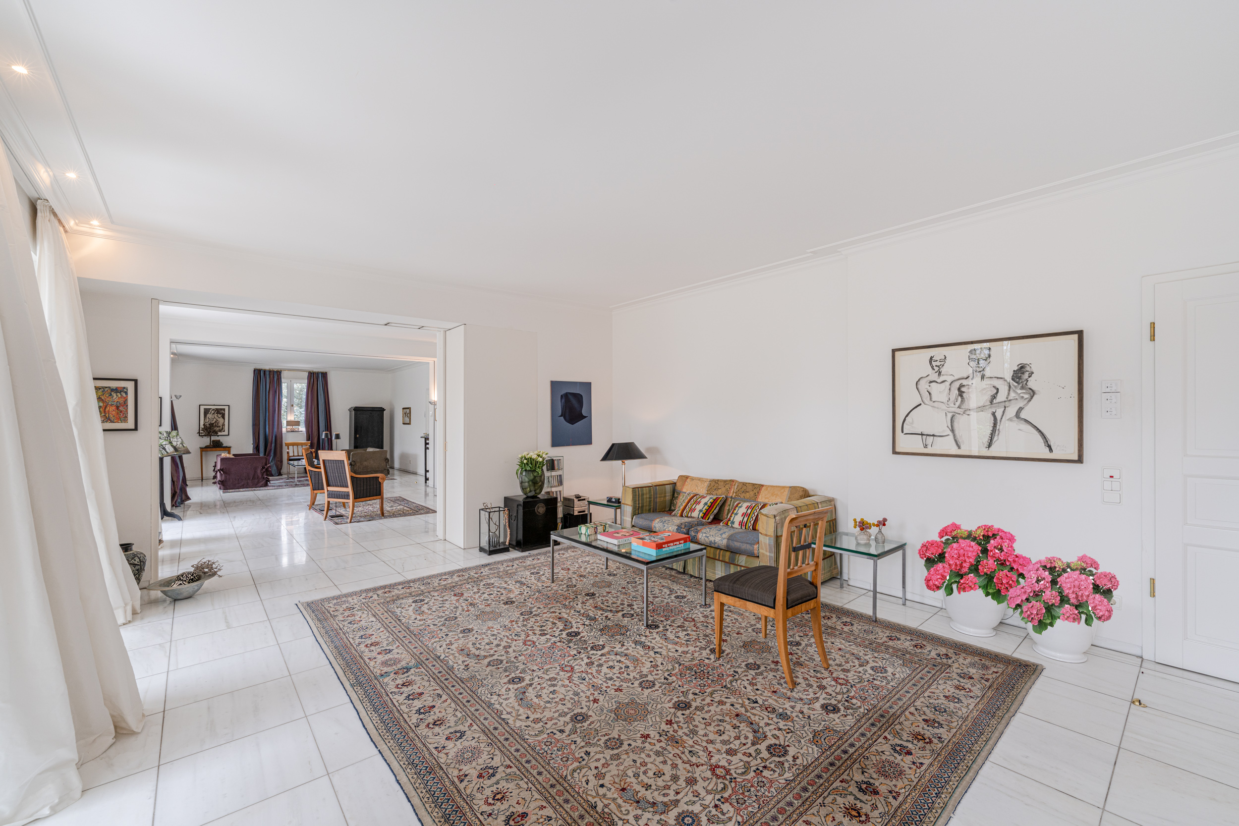 Helles Wohnzimmer mit Teppich, Sofa, Glastisch, Stuhl und rosa Blumen in Töpfen auf Fliesenboden.