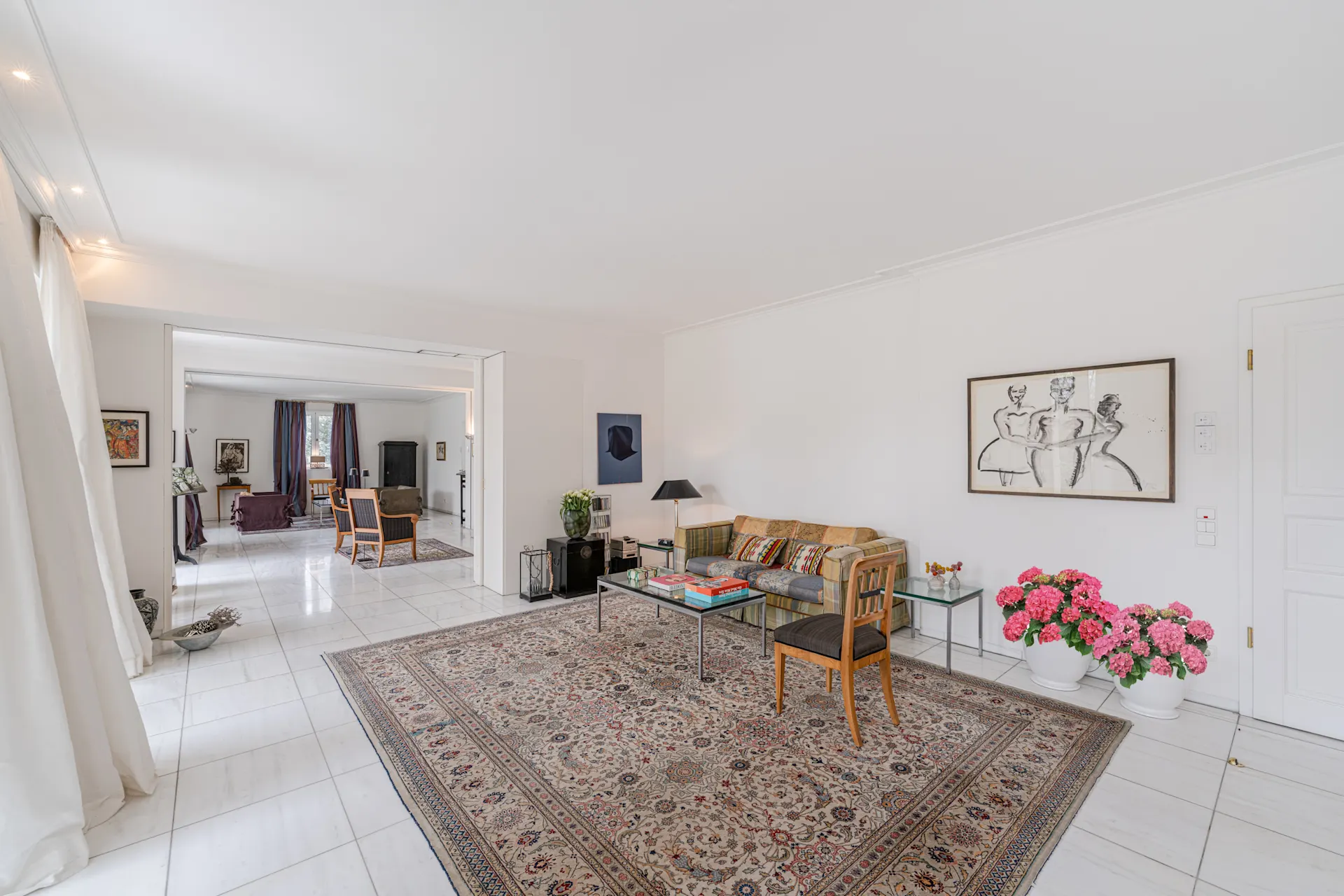 Helles Wohnzimmer mit Teppich, Sofa, Glastisch, Stuhl und rosa Blumen in Töpfen auf Fliesenboden.