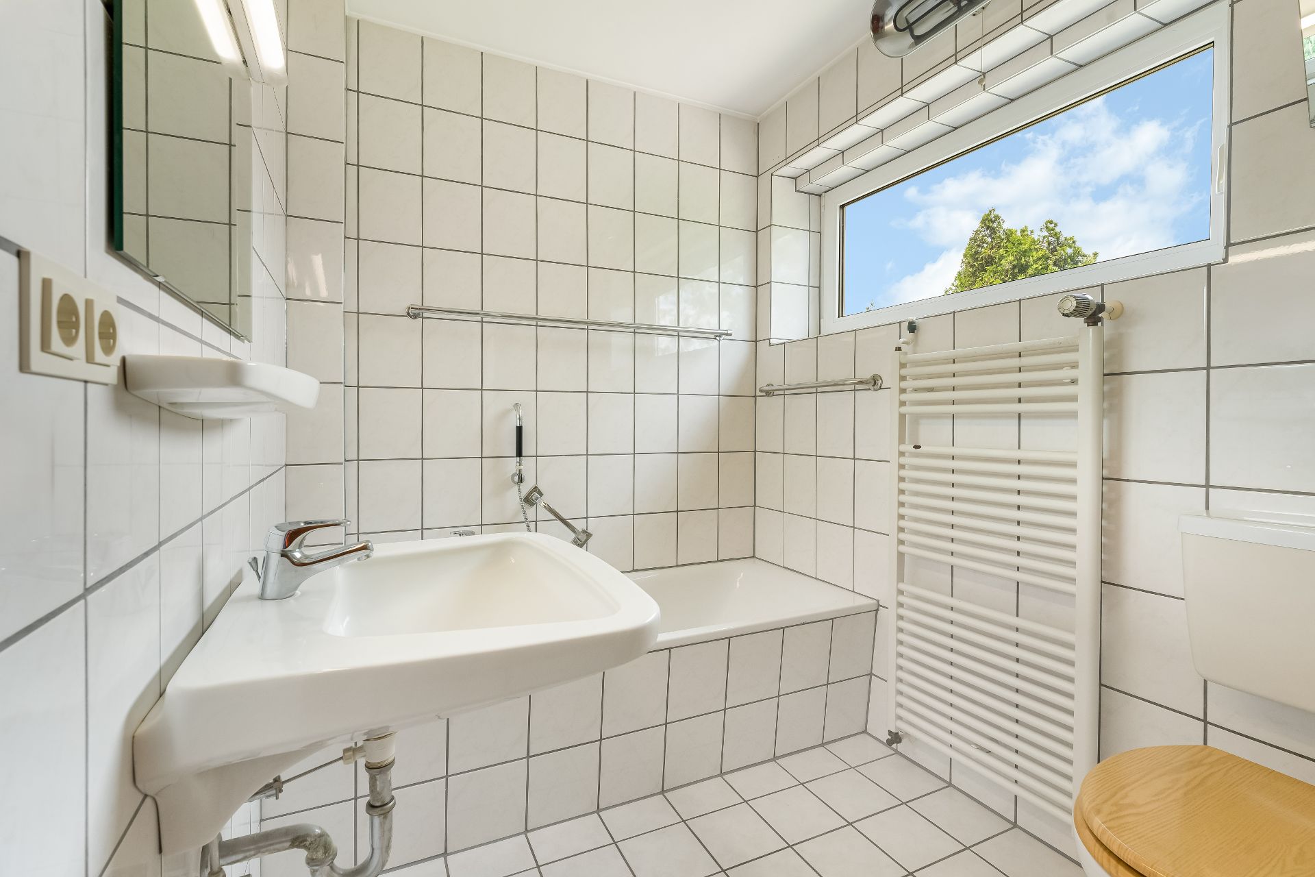 Helles Badezimmer mit weiß gefliesten Wänden, Waschbecken, Badewanne, Heizkörper und Fenster.