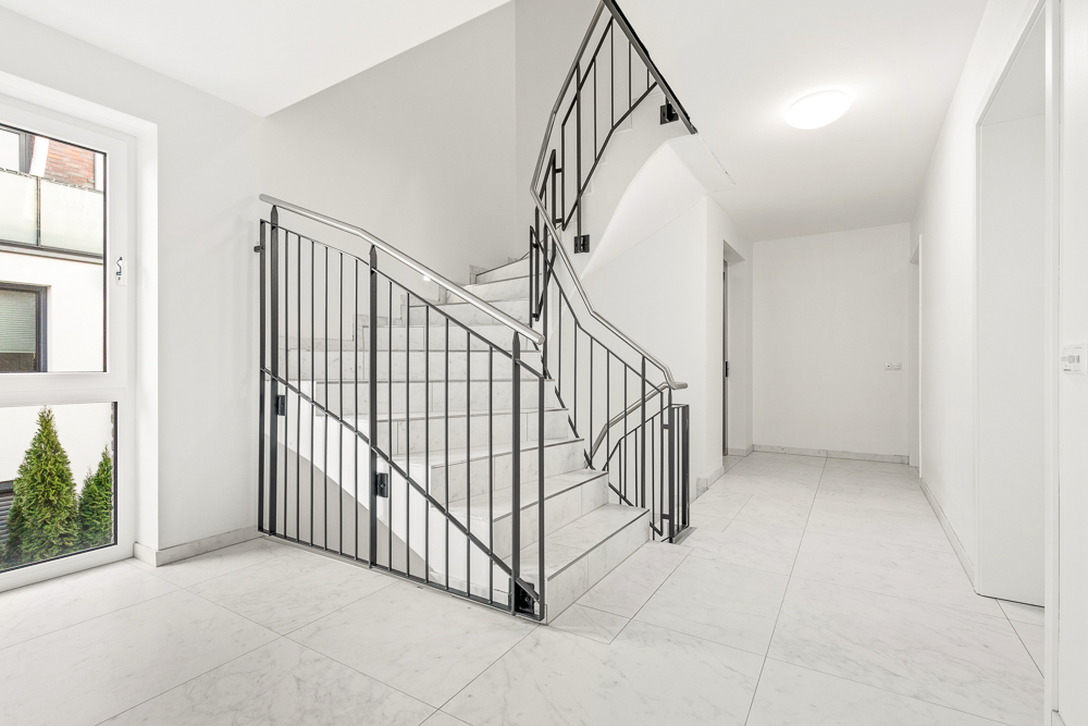 Heller Flur mit Marmortreppe und schwarzem Geländer, große Fenster und weiße Wände.