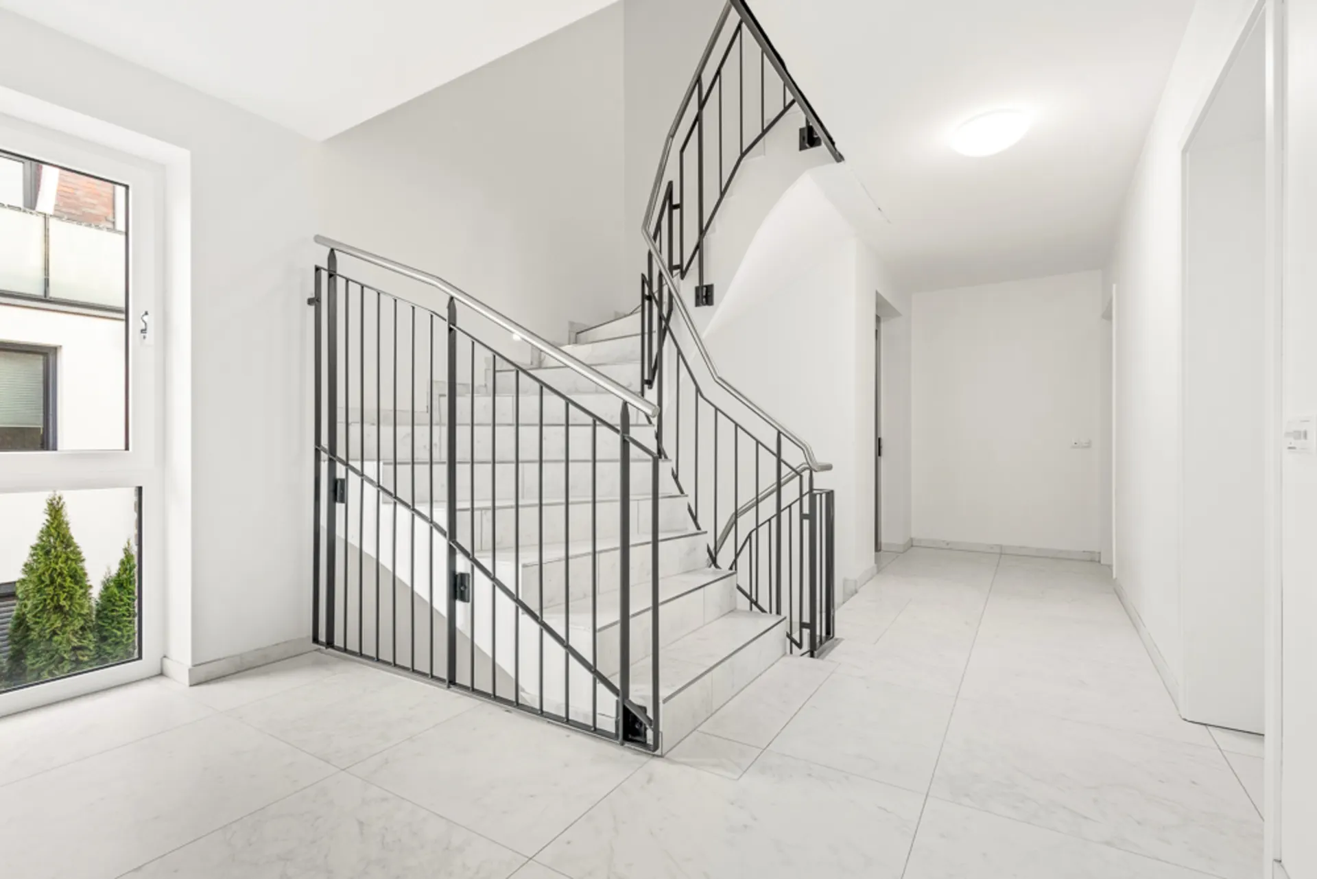 Heller Flur mit Marmortreppe und schwarzem Geländer, große Fenster und weiße Wände.