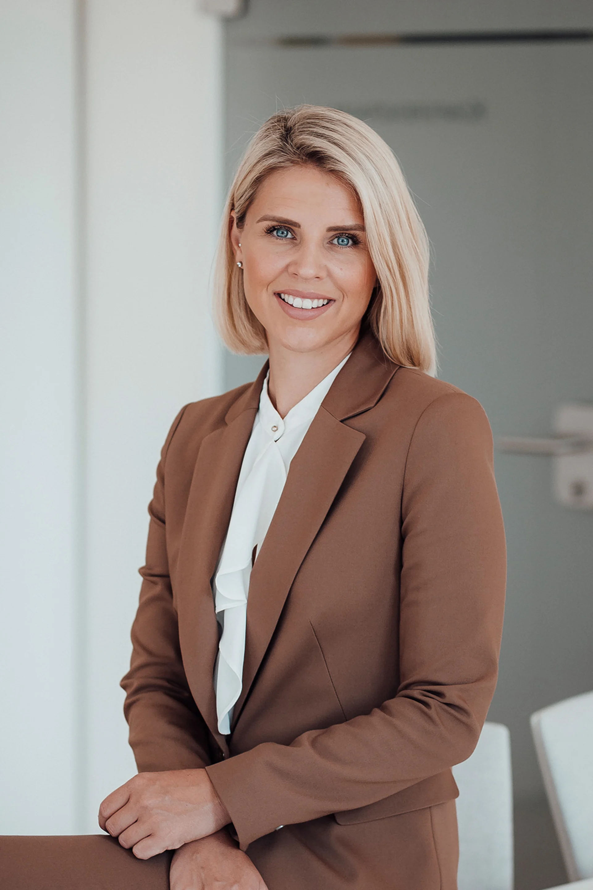 Frau mit blondem Haar im braunen Blazer sitzt und lächelt in modernem, hellen Raum.