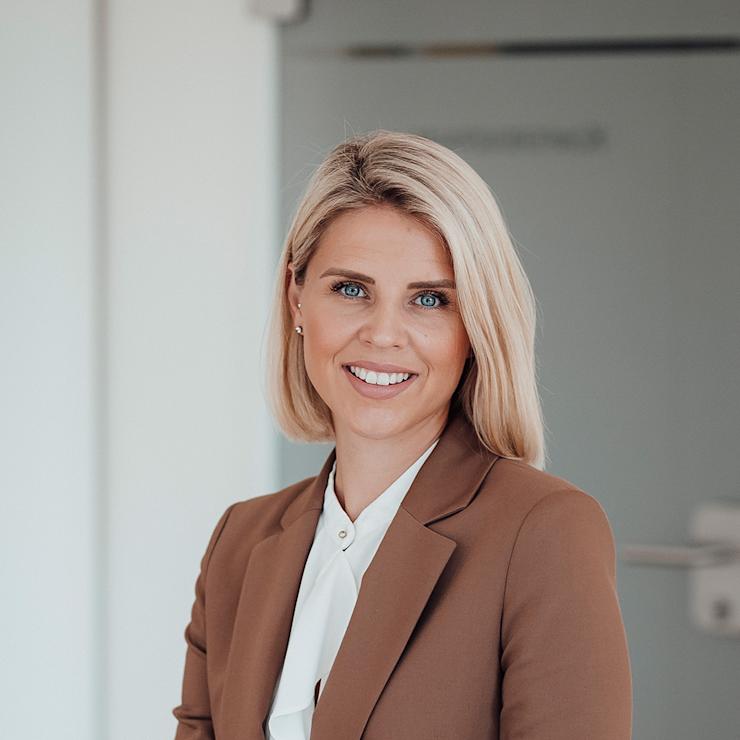 Frau mit blondem Haar im braunen Blazer sitzt und lächelt in modernem, hellen Raum.