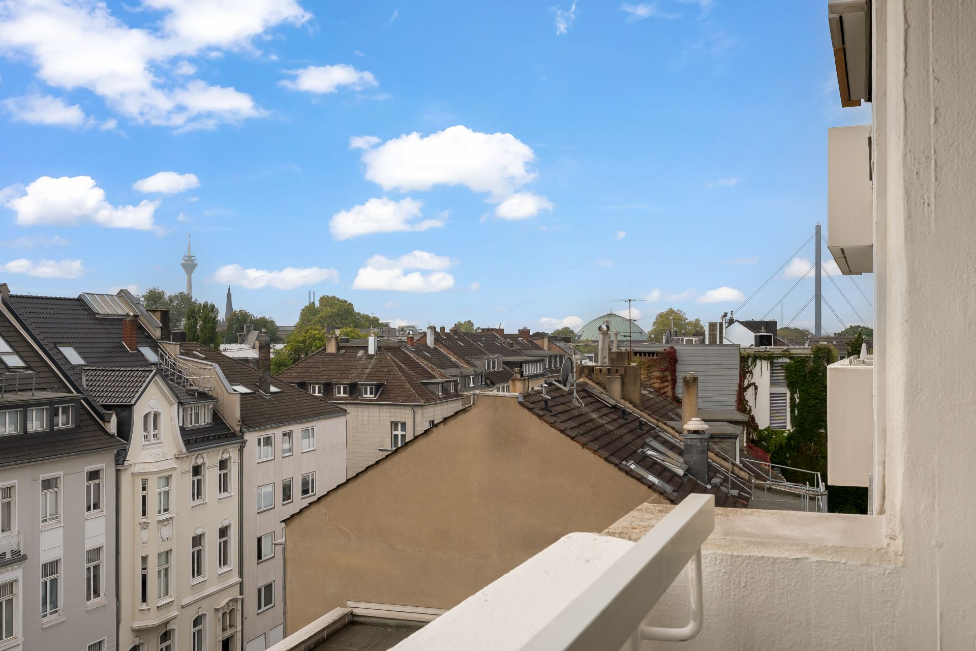 Blick von einem Balkon auf Dächer und Häuser einer Wohngegend bei blauem Himmel.