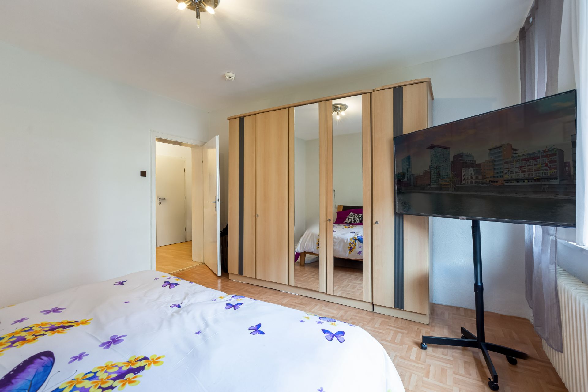 Schlafzimmer mit Doppelbett, großem Kleiderschrank mit Spiegel und Fernseher auf Ständer am Fenster.