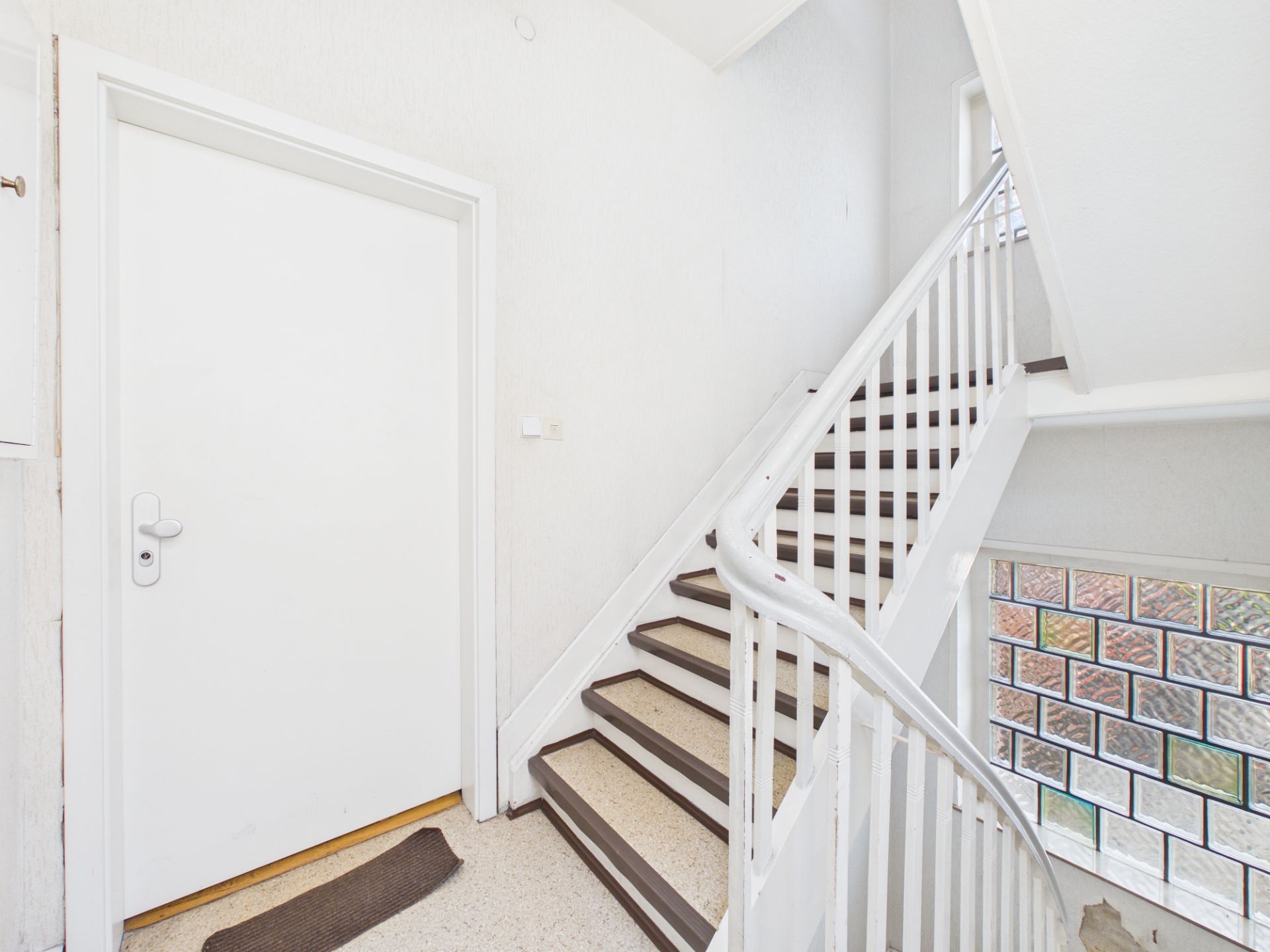 Heller Treppenhausbereich mit weißer Tür, Treppe und Glasbausteinfenster.