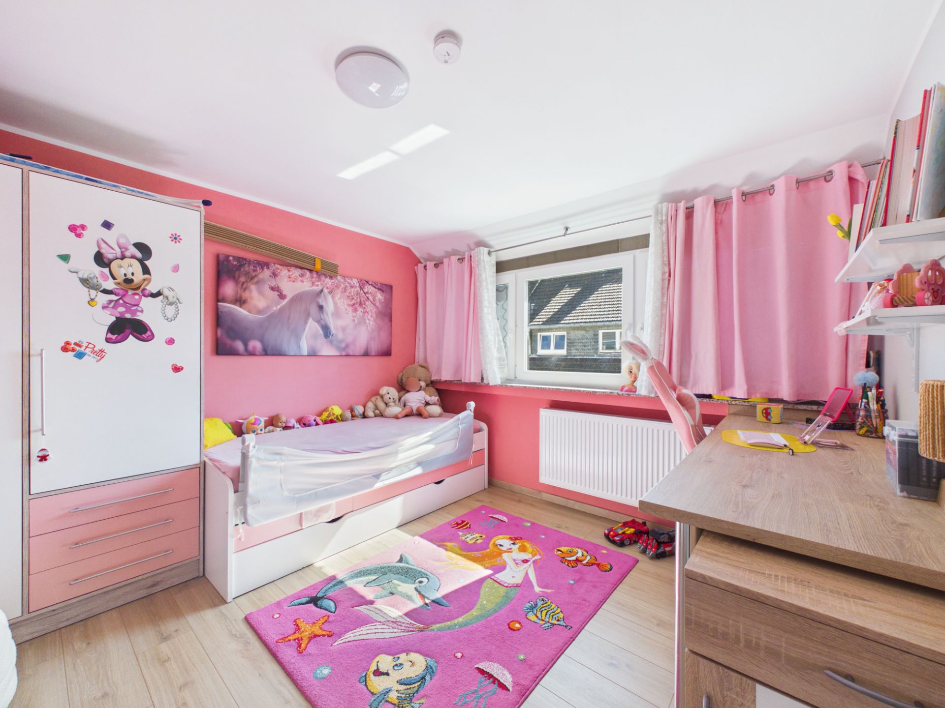 Helles Kinderzimmer mit rosa Wänden, Spielteppich, Bett, Schreibtisch und Fenster mit pinken Vorhängen.