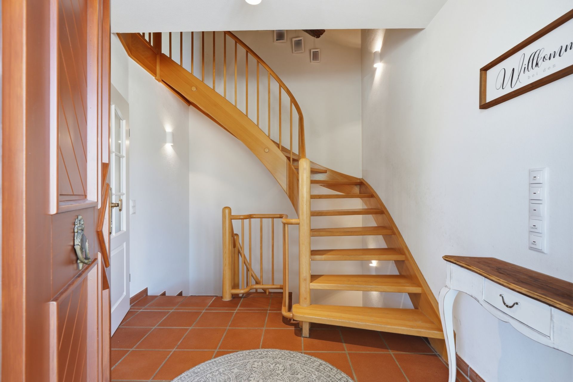 Heller Eingangsbereich mit hölzerner Treppe, Fliesenboden und kleinem Tisch an der Wand.