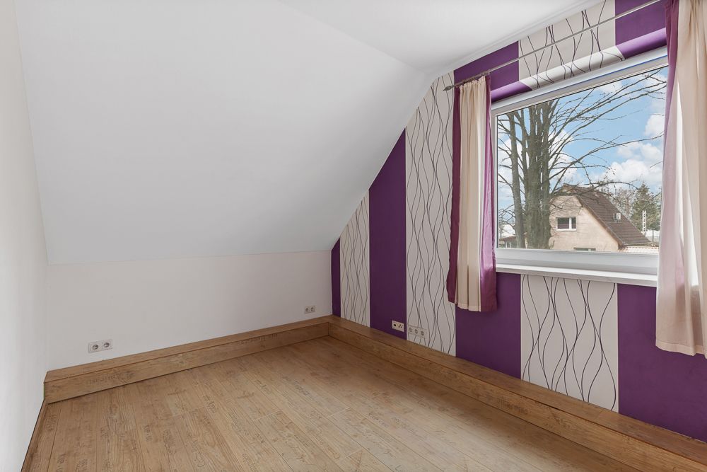 Leeres Zimmer mit schräger Decke, lila-gemusterter Tapete und großem Fenster mit Vorhängen.