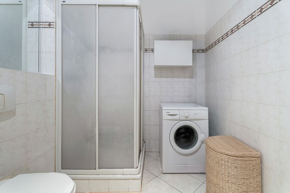 Badezimmer mit Duschkabine, Waschmaschine und Wäschekorb auf gefliestem Boden.