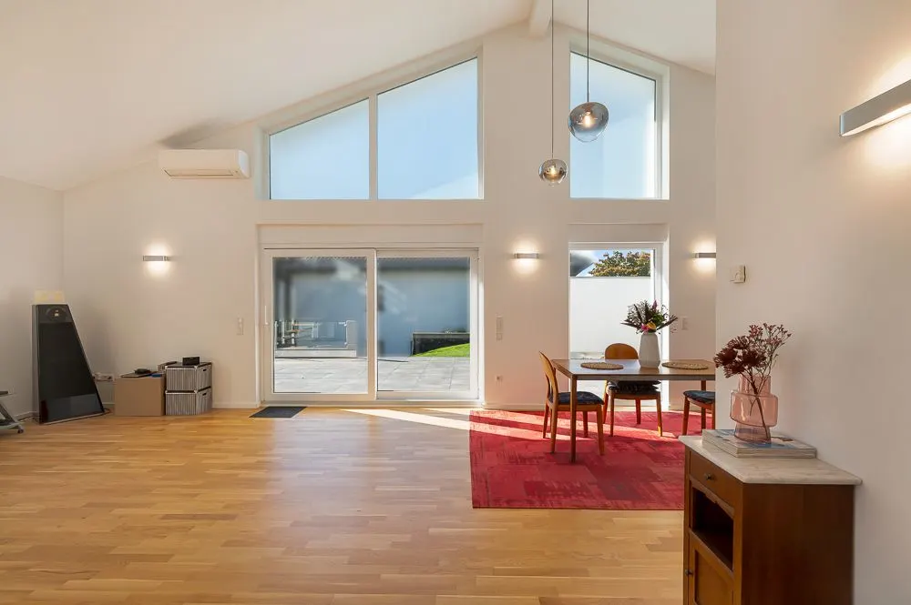 Helles Wohnzimmer mit Holzboden, Esstisch auf rotem Teppich und großen Fenstern zur Terrasse.