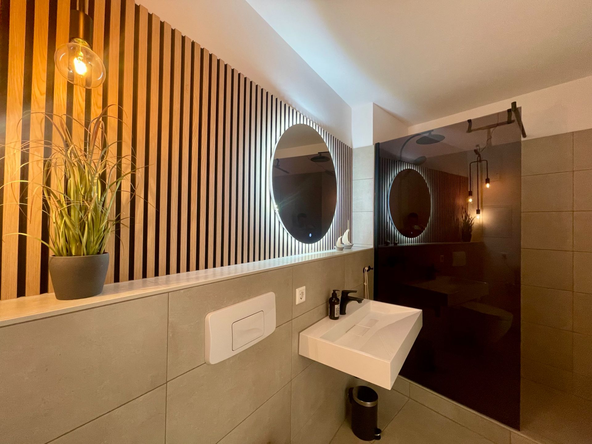 Modernes Badezimmer mit Waschbecken, rundem Spiegel, Holzverkleidung und Glasdusche.
