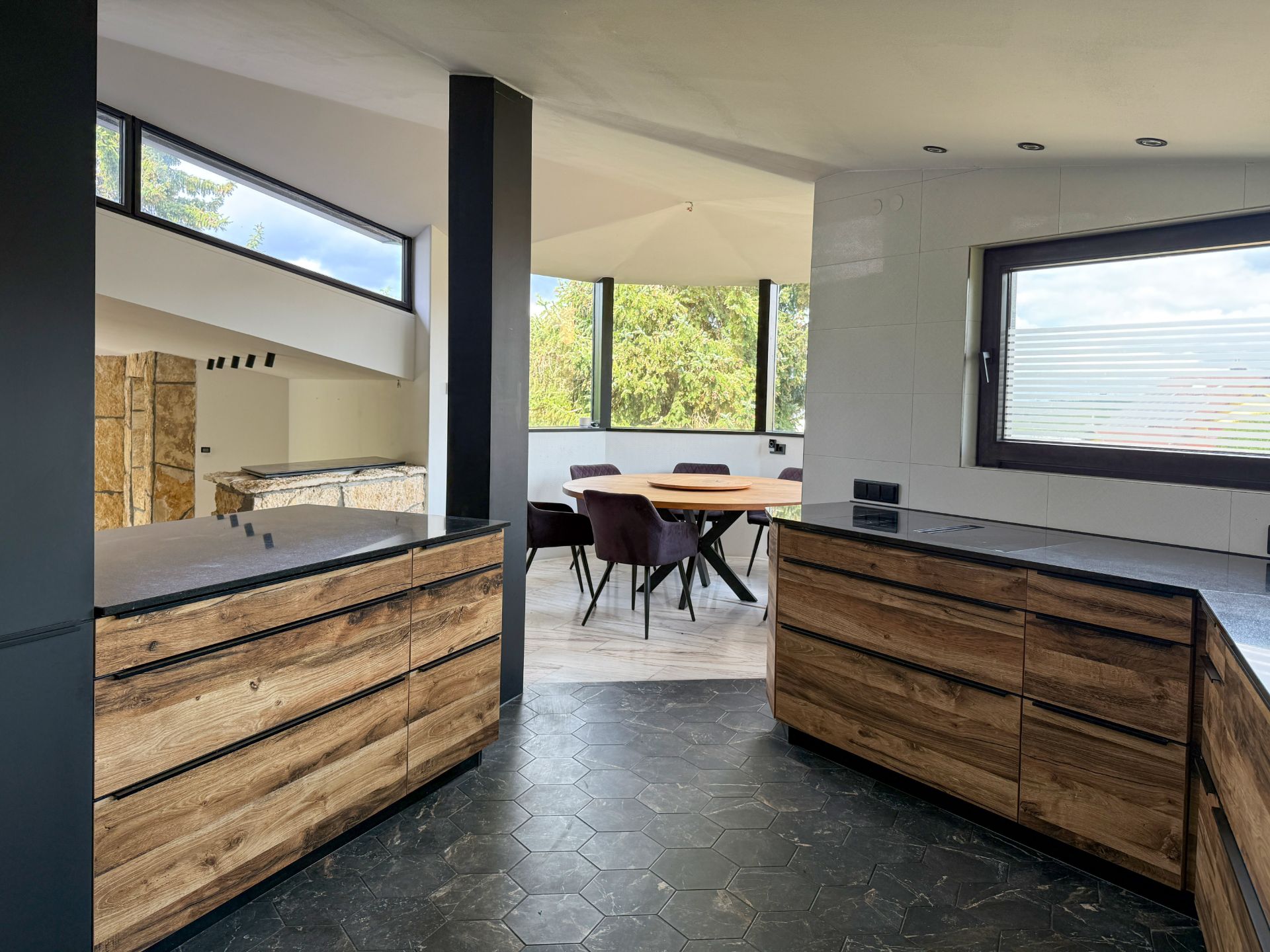 Moderne Küche mit Holzfronten und schwarzem Boden, dahinter ein Essbereich mit großem Fenster.