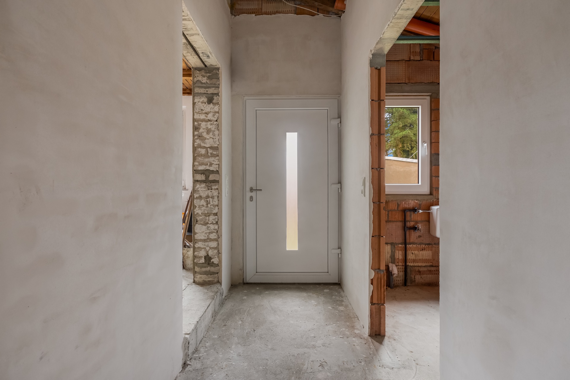 Flur mit weißer Eingangstür, sichtbaren Rohbauwänden und Fenster auf der rechten Seite.