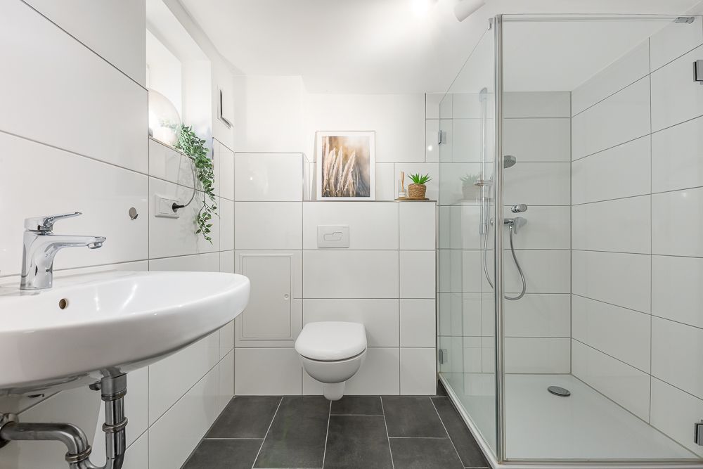 Modernes Bad mit Waschbecken, Hänge-WC und Glasdusche, weiße Fliesen und Pflanzen.