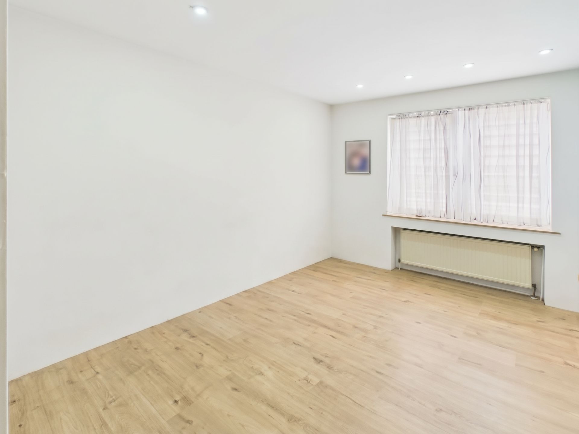 Leerer Raum mit hellem Holzboden, weißer Wand und großem Fenster mit Vorhang.