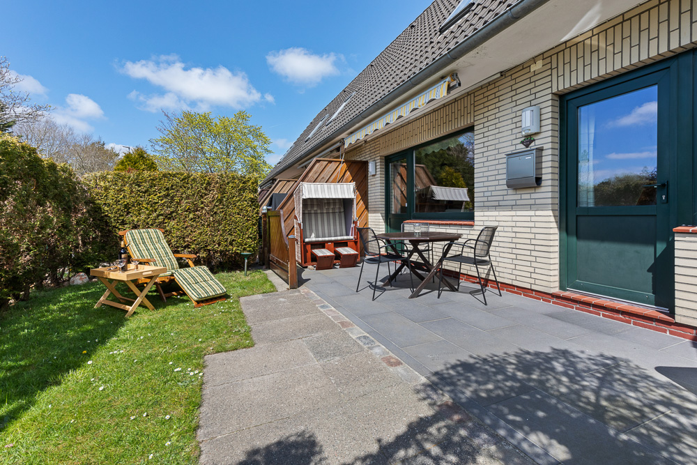 Terrasse mit Tisch, Stühlen, Liegestühlen und Strandkorb auf Rasen vor einem Haus.