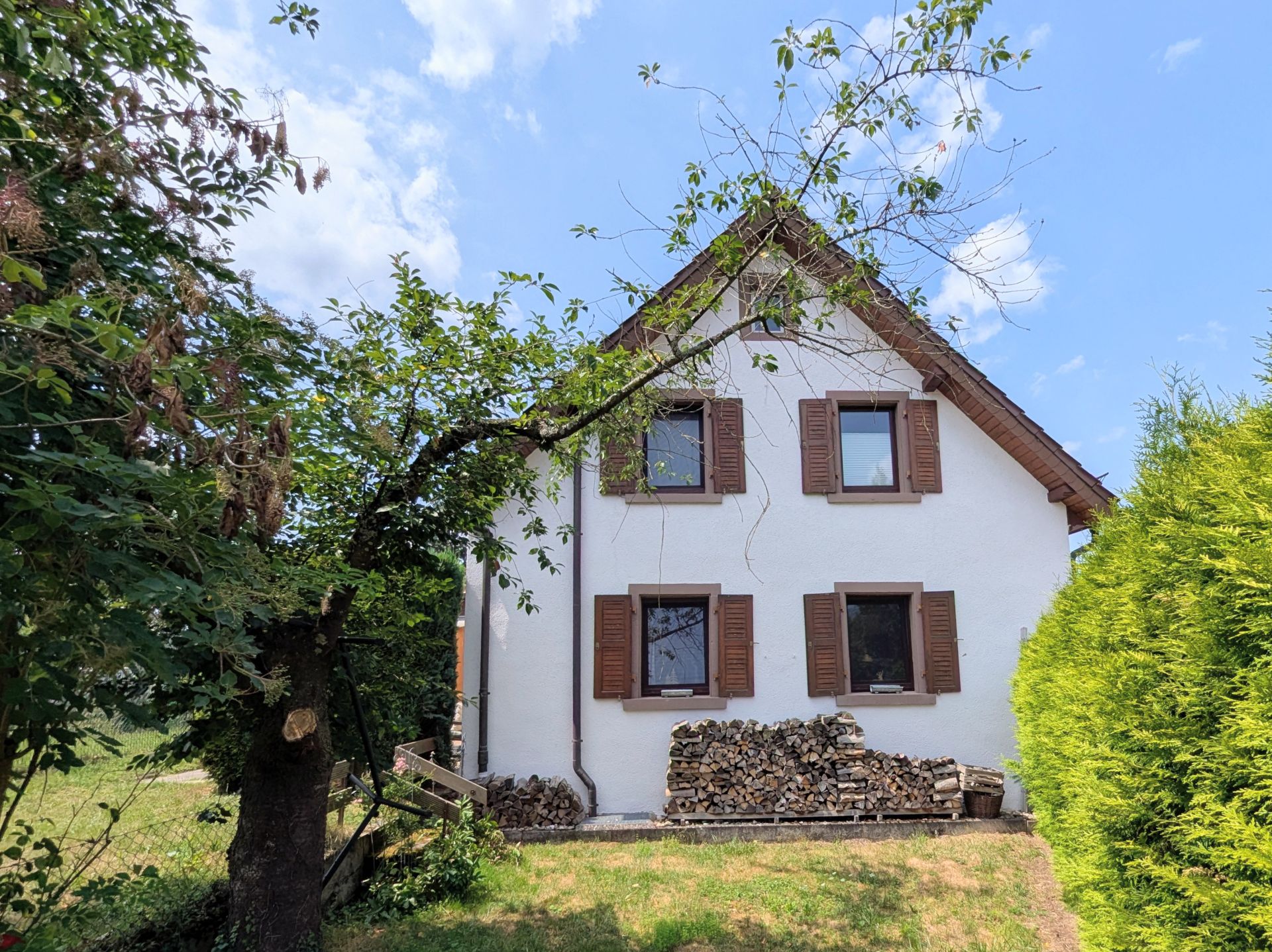 Weißes Haus mit braunen Fensterläden und gestapeltem Brennholz im Garten.