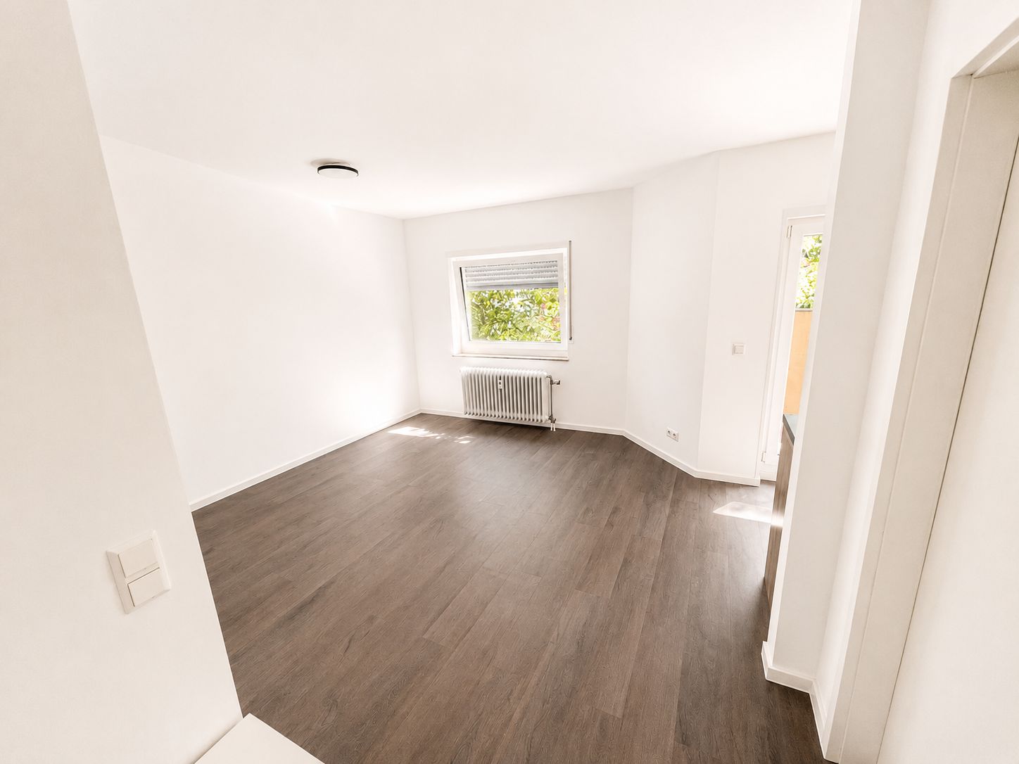 Heller, leerer Raum mit dunklem Boden, weißer Wand, Fenster und Heizkörper.