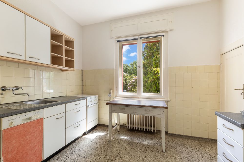 Helle Küche mit weißen Schränken, großem Fenster und einfachem Tisch vor der Heizung.