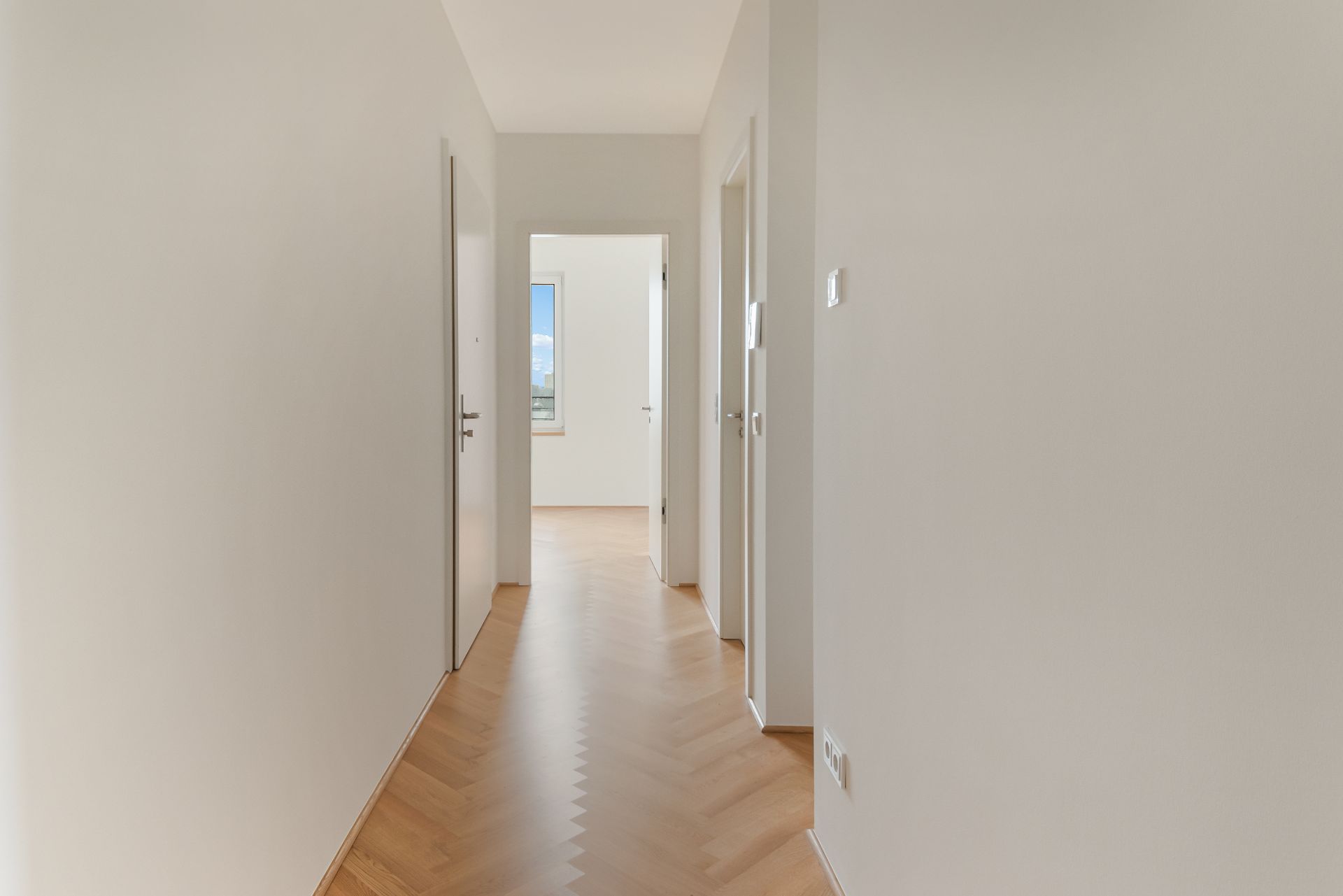 Heller Flur mit Holzboden und mehreren offenen Türen, am Ende ein Fenster mit Ausblick.