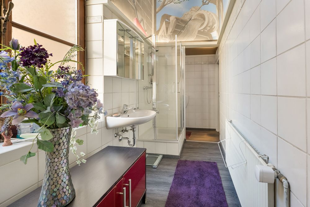 Modernes Bad mit Dusche, Waschbecken, lila Teppich und Blumenstrauß auf einer Kommode.