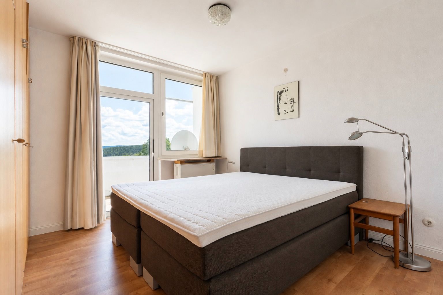 Helles Schlafzimmer mit Doppelbett, großem Fenster, Vorhängen und Nachttisch mit Lampe.