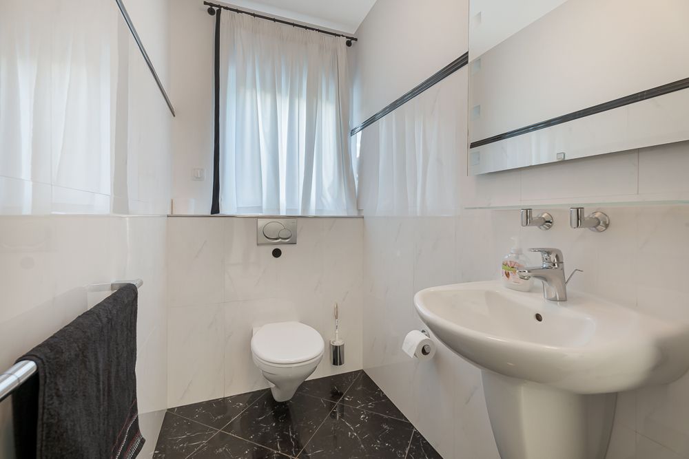 Helles Badezimmer mit WC, Waschbecken, Spiegel und Fenster mit weißen Vorhängen.