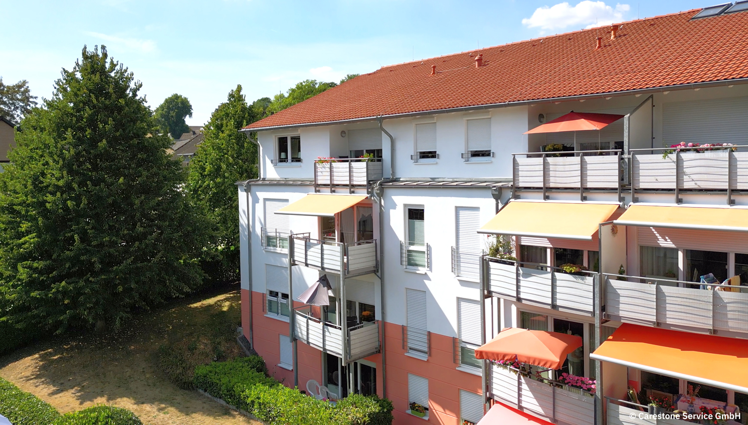 Mehrfamilienhaus mit weißen und orangen Markisen und mehreren Balkonen, umgeben von Bäumen.