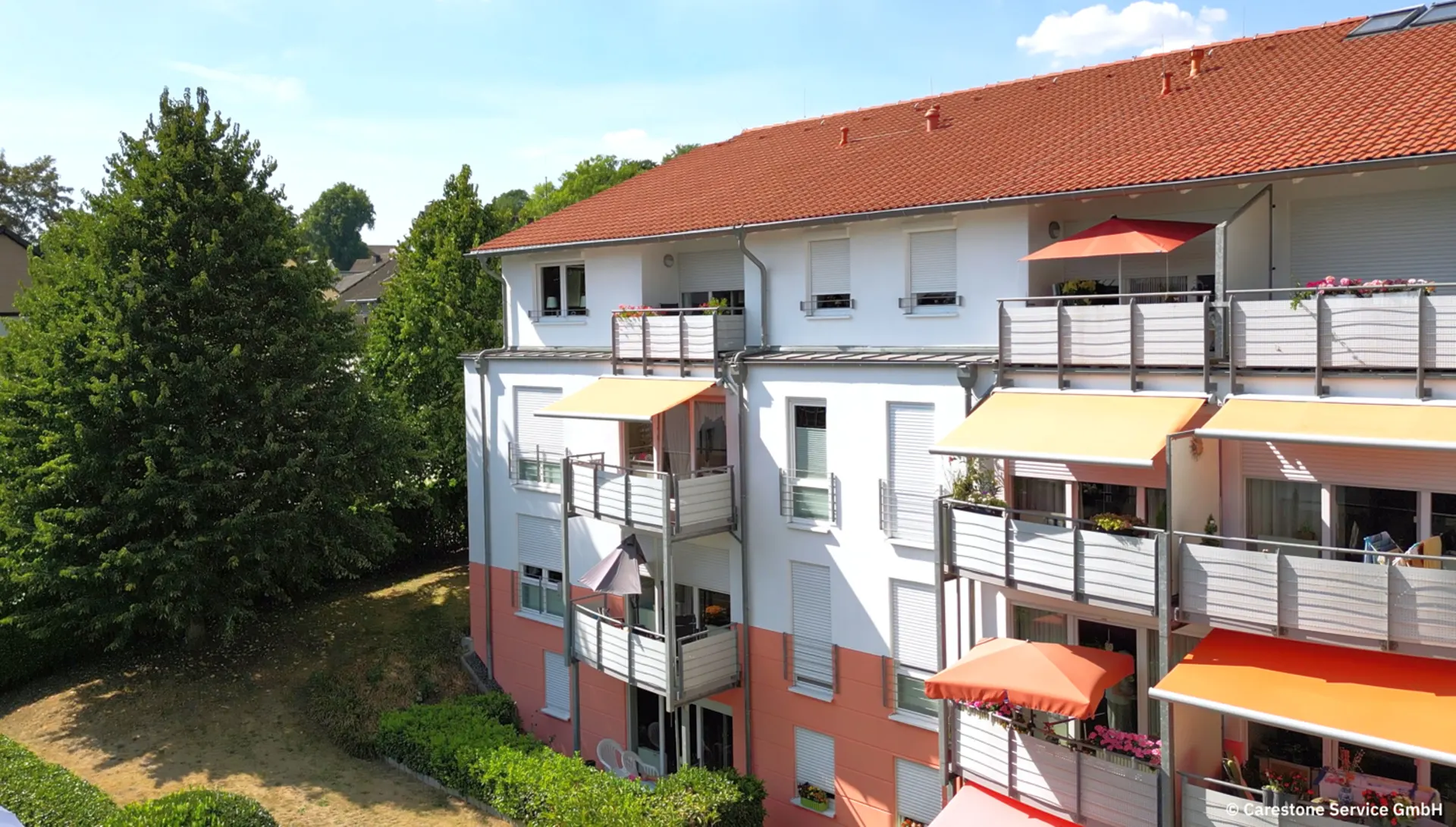 Mehrfamilienhaus mit weißen und orangen Markisen und mehreren Balkonen, umgeben von Bäumen.