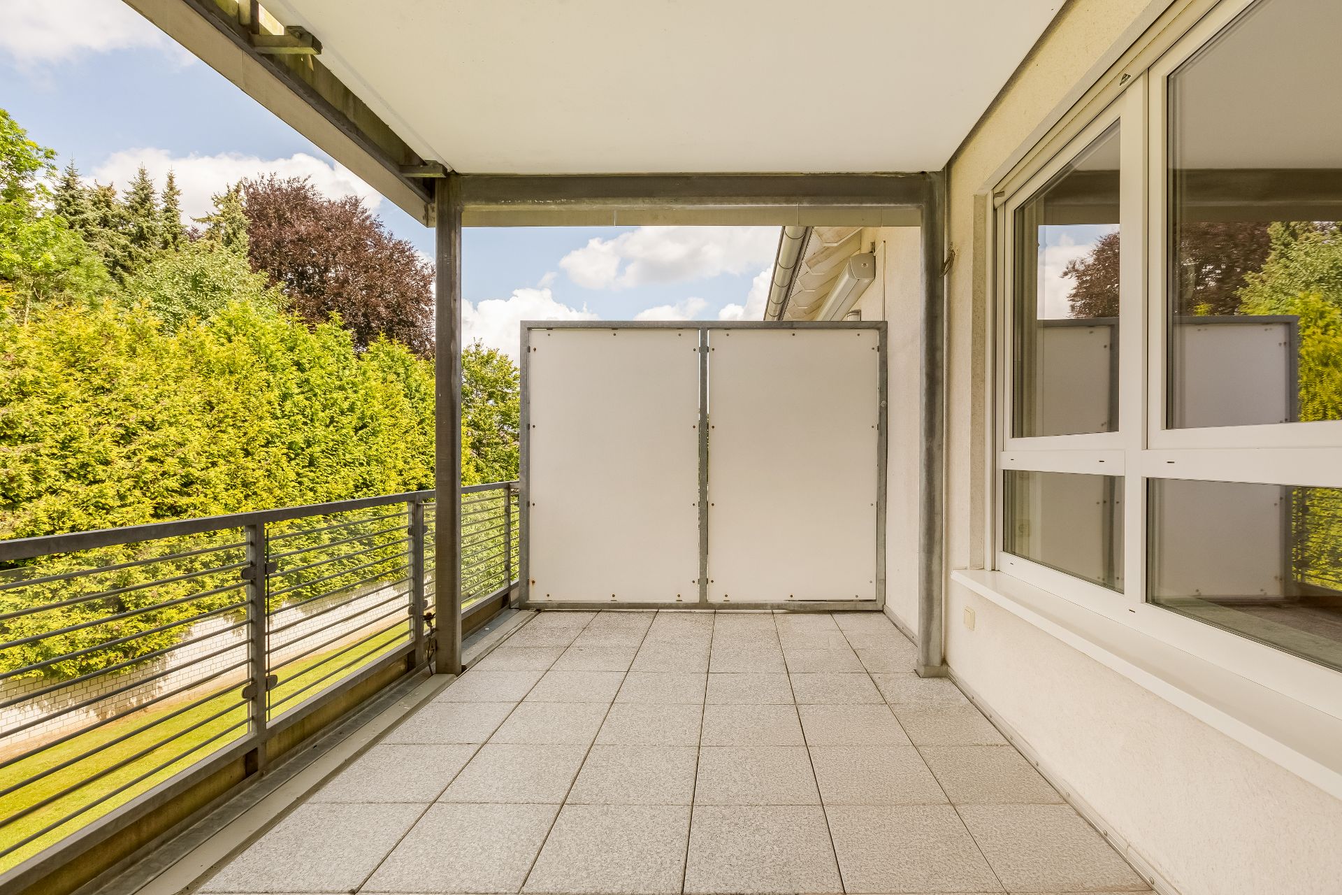Überdachter Balkon mit Fliesenboden, Geländer und Sichtschutz, Blick auf grüne Bäume.