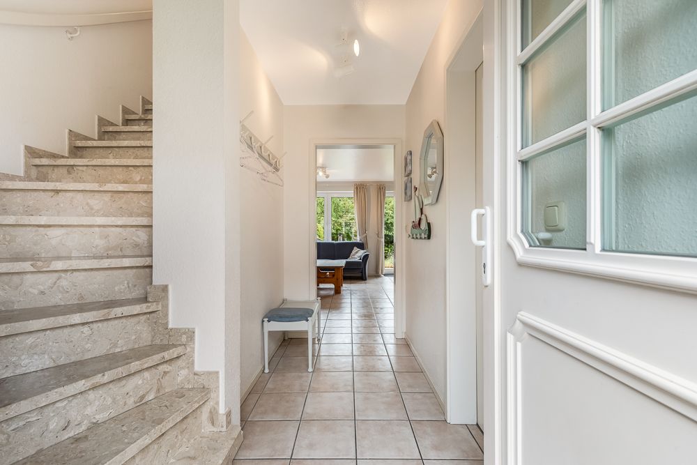 Heller Flur mit Fliesenboden, Treppe links und Blick ins Wohnzimmer mit Sofa.