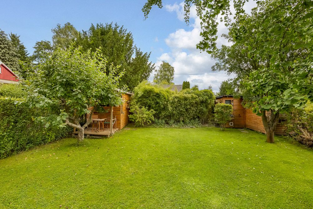 Grüner Garten mit zwei Holzhütten, Bäumen und einer kleinen Terrasse mit Sitzmöbeln.